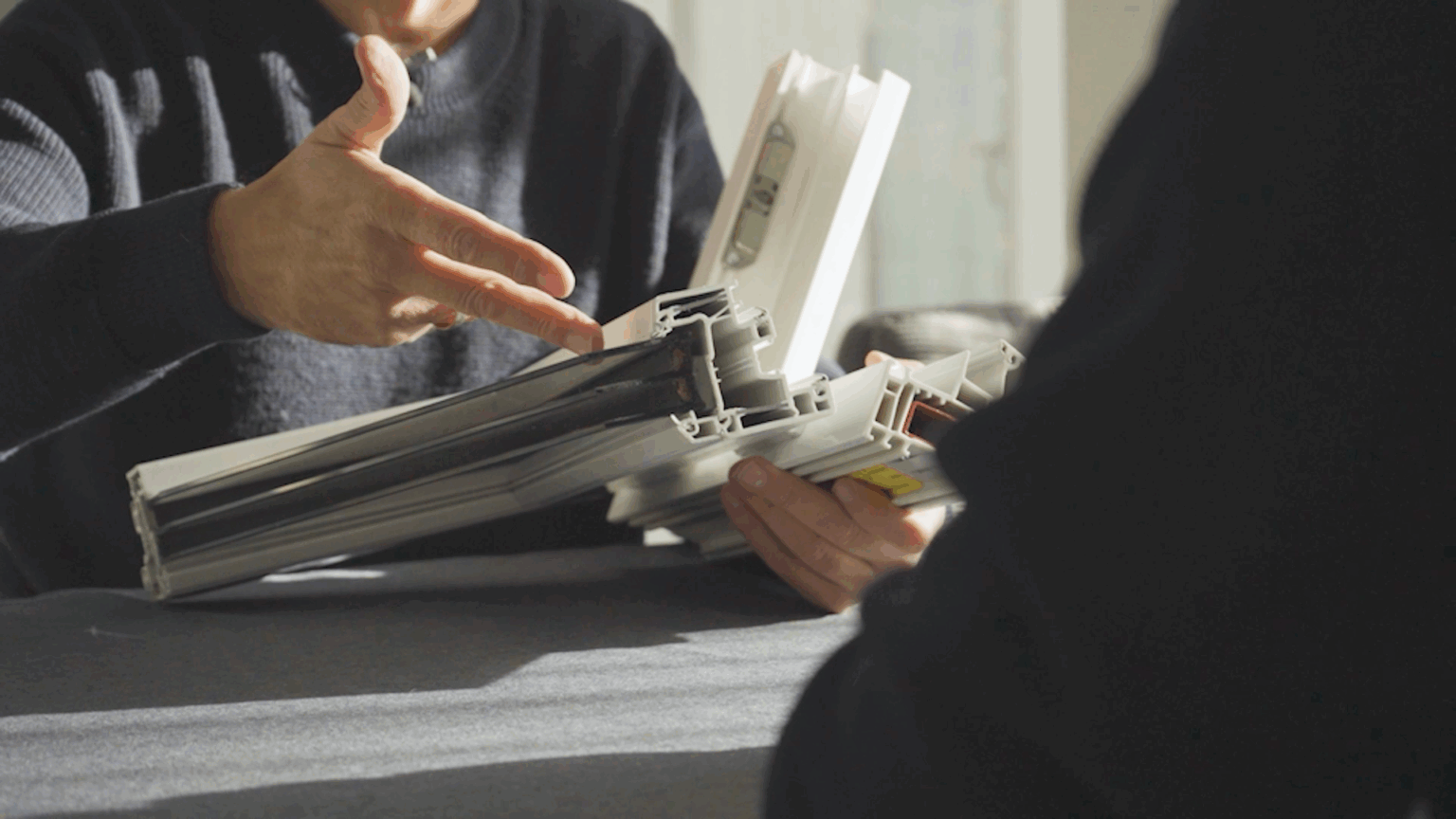 Beratung-2 Person zeigt PVC-Fensterprofil in Nahaufnahme, erklärt Isolierung und Aufbau bei Tageslicht.