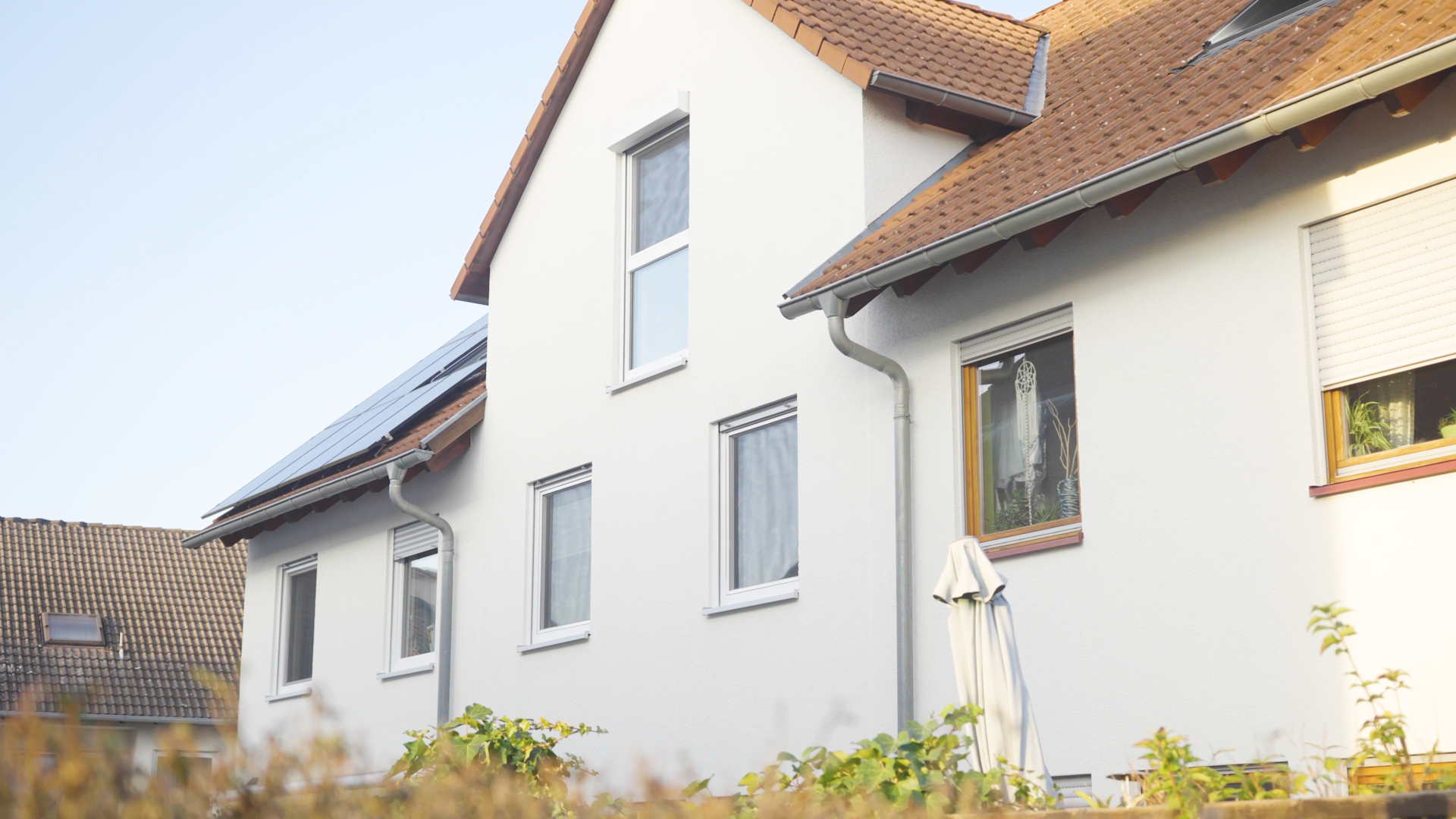 Fenster-20 Mehrfamilienhaus mit Solarpanel auf dem Dach, umgeben von Pflanzen und blauer Himmel im Hintergrund.