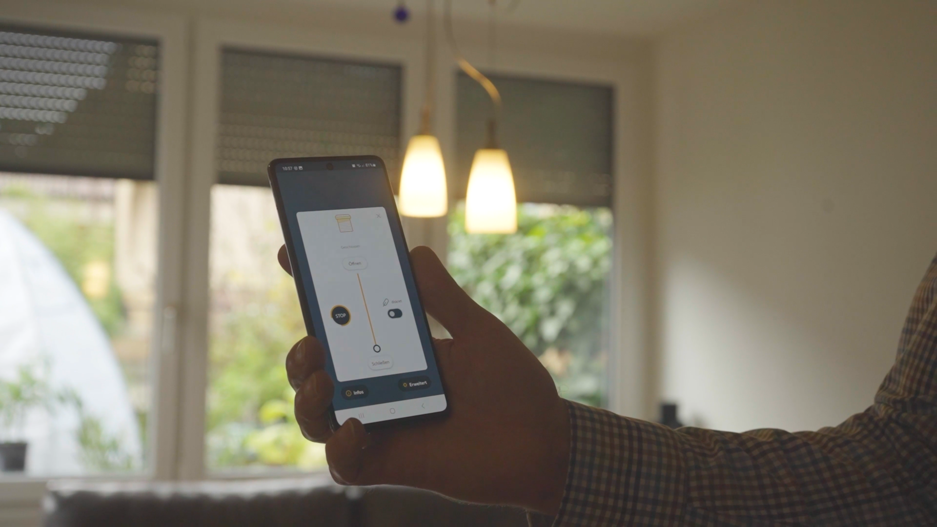 Rolladen-8 Person steuert smarte Jalousien-App auf Smartphone im Wohnzimmer. Im Hintergrund sind Fenster und Lampen sichtbar.
