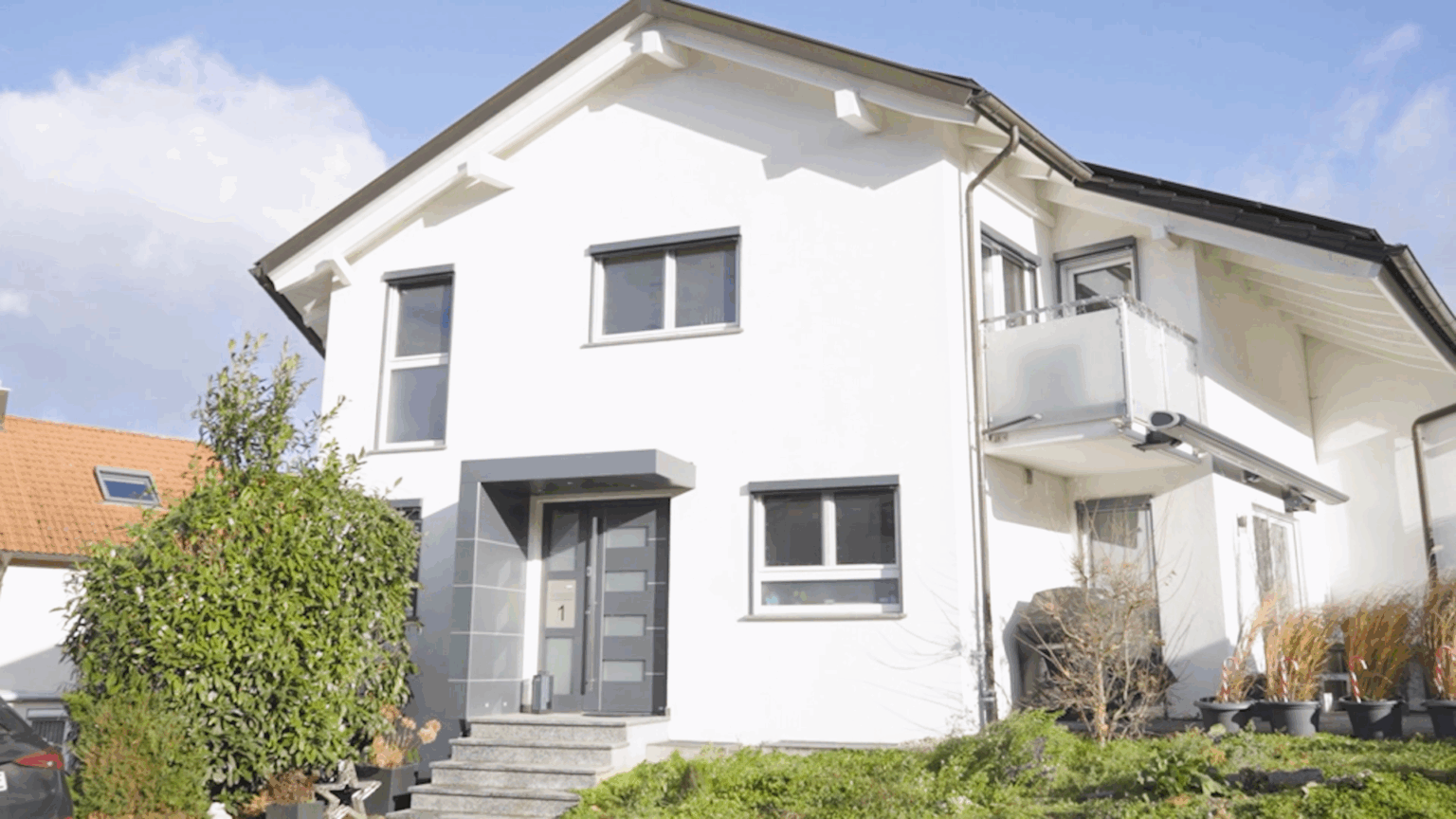 Türen-10 Moderne Doppelhaushälfte mit weißer Fassade und Balkon an sonnigem Tag, umgeben von grüner Bepflanzung.